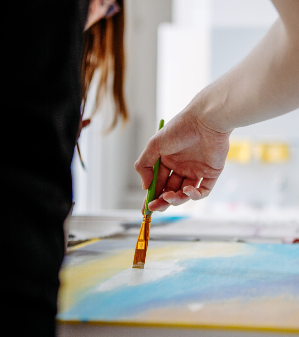 Eine Hand hält einen Pinsel und streicht mit weißer Farbe über ein Blatt Papier, das mit etwas blauer Farbe bemalt ist.