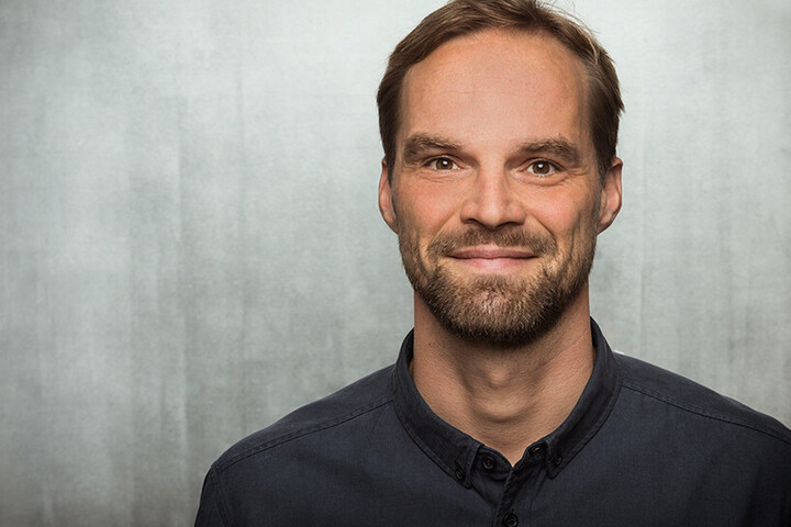 Robert Bach lächelt in die Kamera. Er trägt ein dunkles Hemd und steht vor einer grauen Wand.